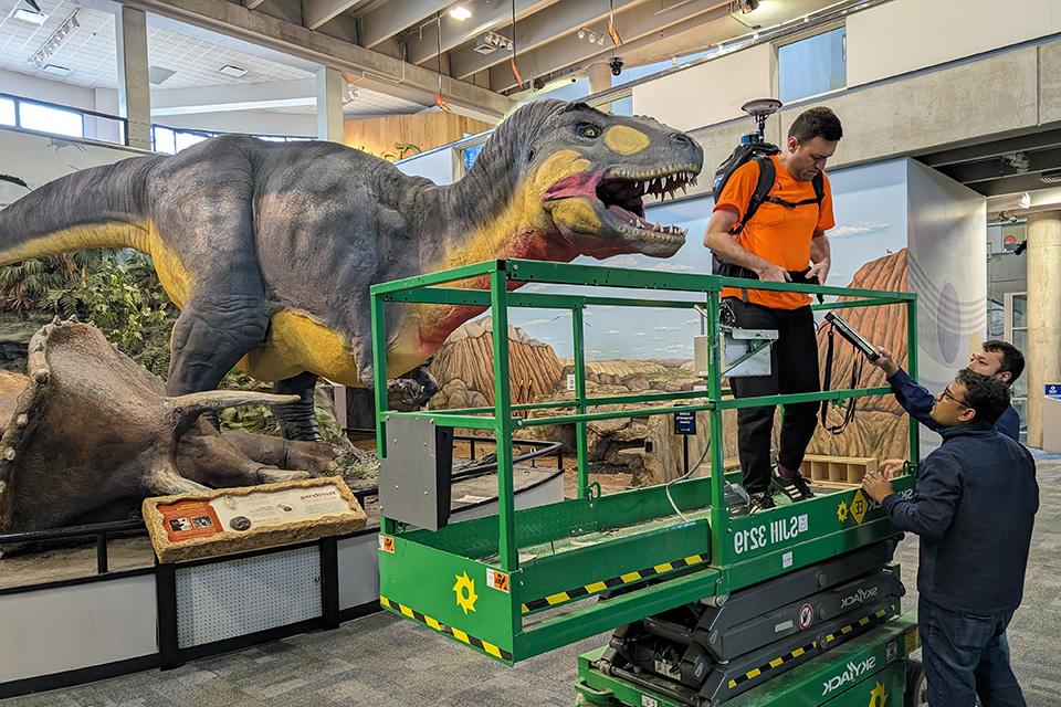 A student stands on a lift with a large piece of equipment on his back in front of one of the latex dinosaurs.