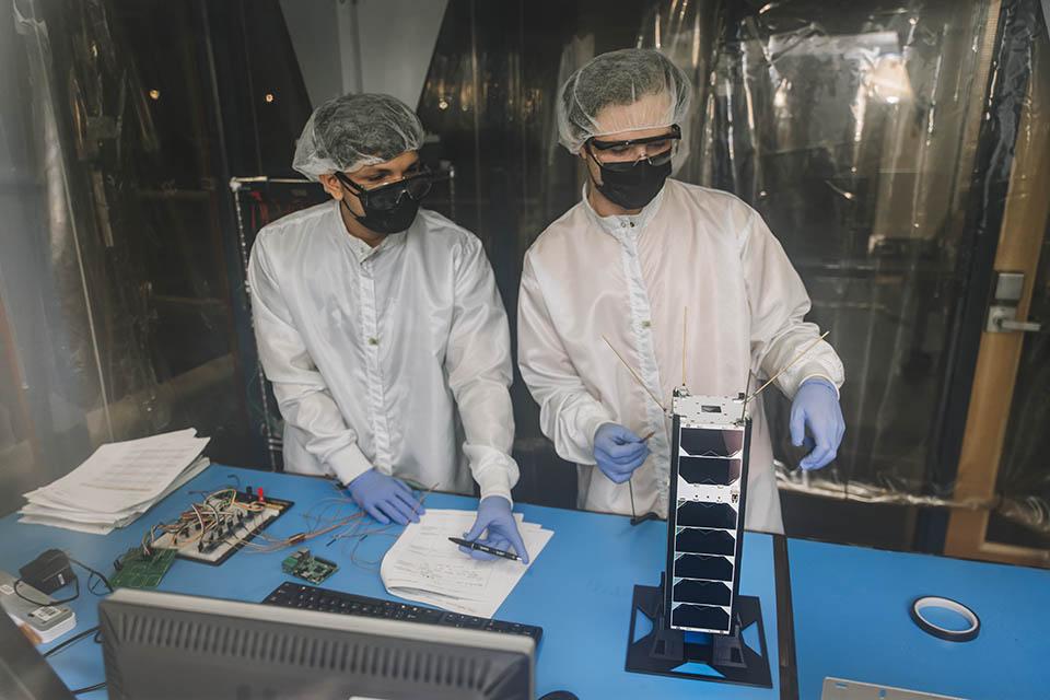 Two students work on a small satellite at a scientific work bench.