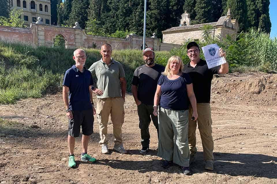 A group of researchers stand near the Italian dig site. One male researcher holds up a sign that reads "Saint Louis University."